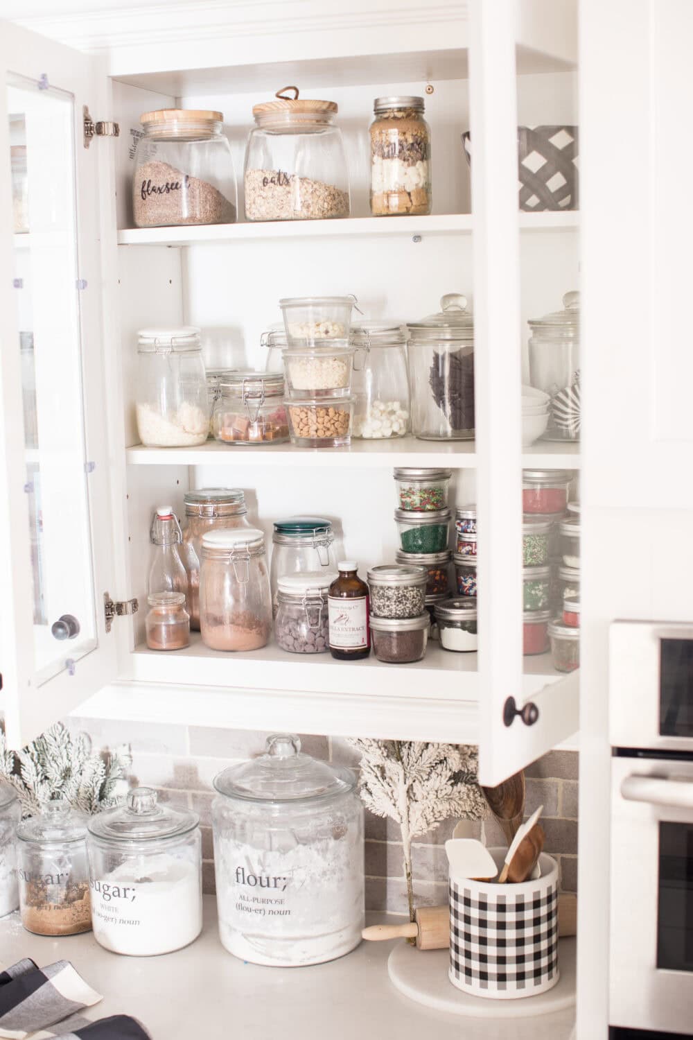 A neatly organized kitchen pantry with glass jars filled with baking ingredients and spices, featuring a white cabinet with open doors for easy access, ideal for food storage and home baking.