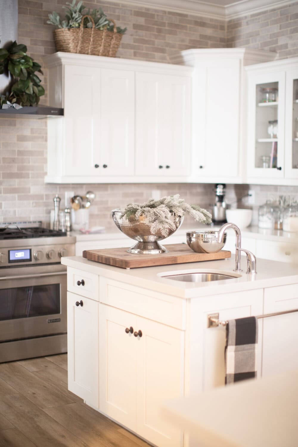 Bright white kitchen cabinetry with a cozy farmhouse style, accented by brushed nickel fixtures and a rustic wooden tray. Elegant silver bowls with greenery add a touch of sophistication to the modern space.